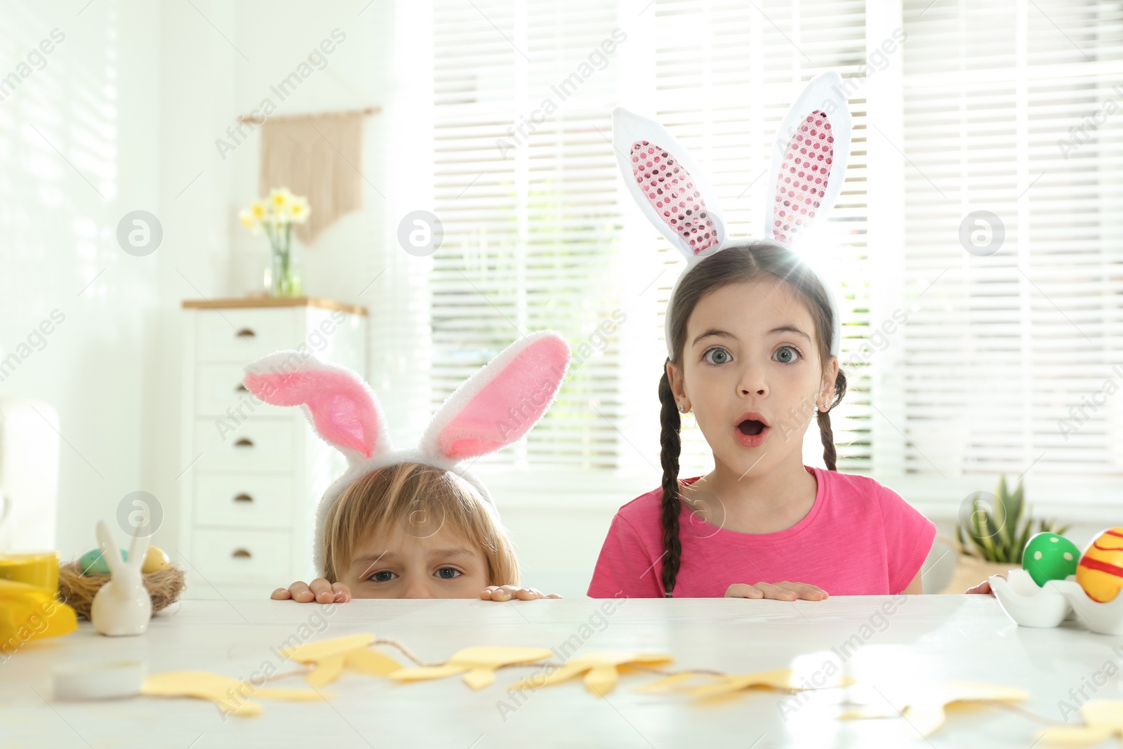 Photo of Emotional children wearing bunny ears headbands at table with Easter eggs, indoors