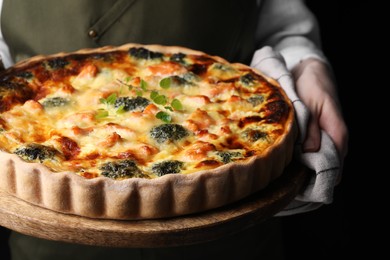 Photo of Woman holding wooden board with delicious homemade salmon quiche on black background, closeup