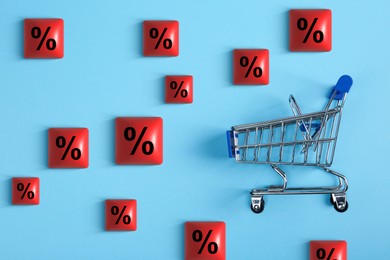Image of Discount offer. Cubes with percent signs and empty shopping trolley on light blue background, flat lay