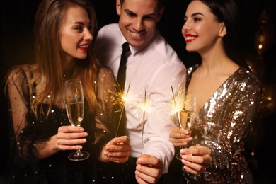 Photo of Happy friends celebrating New Year against black background, focus on hands with glasses of sparkling wine and sparklers