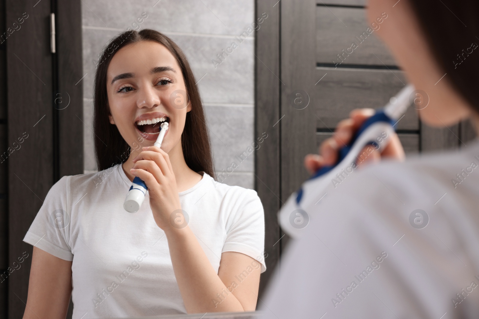 Photo of Young woman brushing her teeth with electric toothbrush near mirror in bathroom