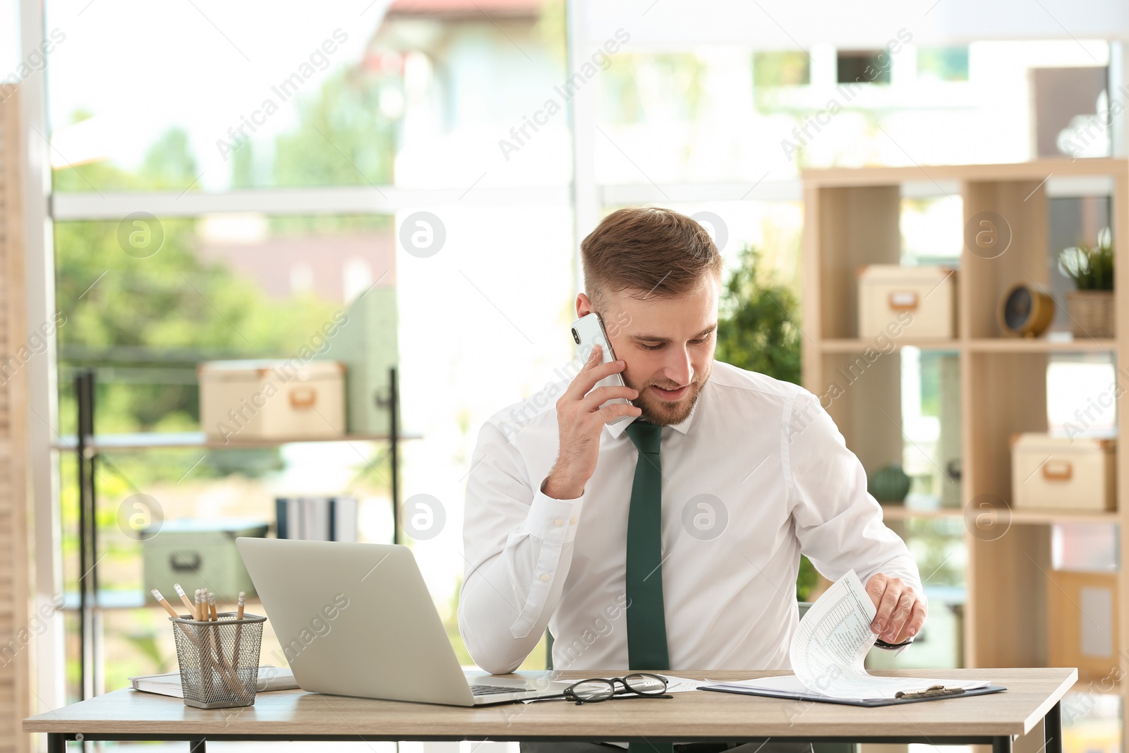 Photo of Young man talking on phone while working with laptop at table in office