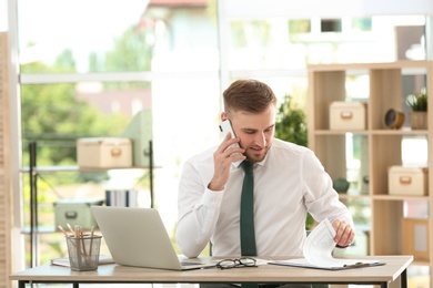 Young man talking on phone while working with laptop at table in office