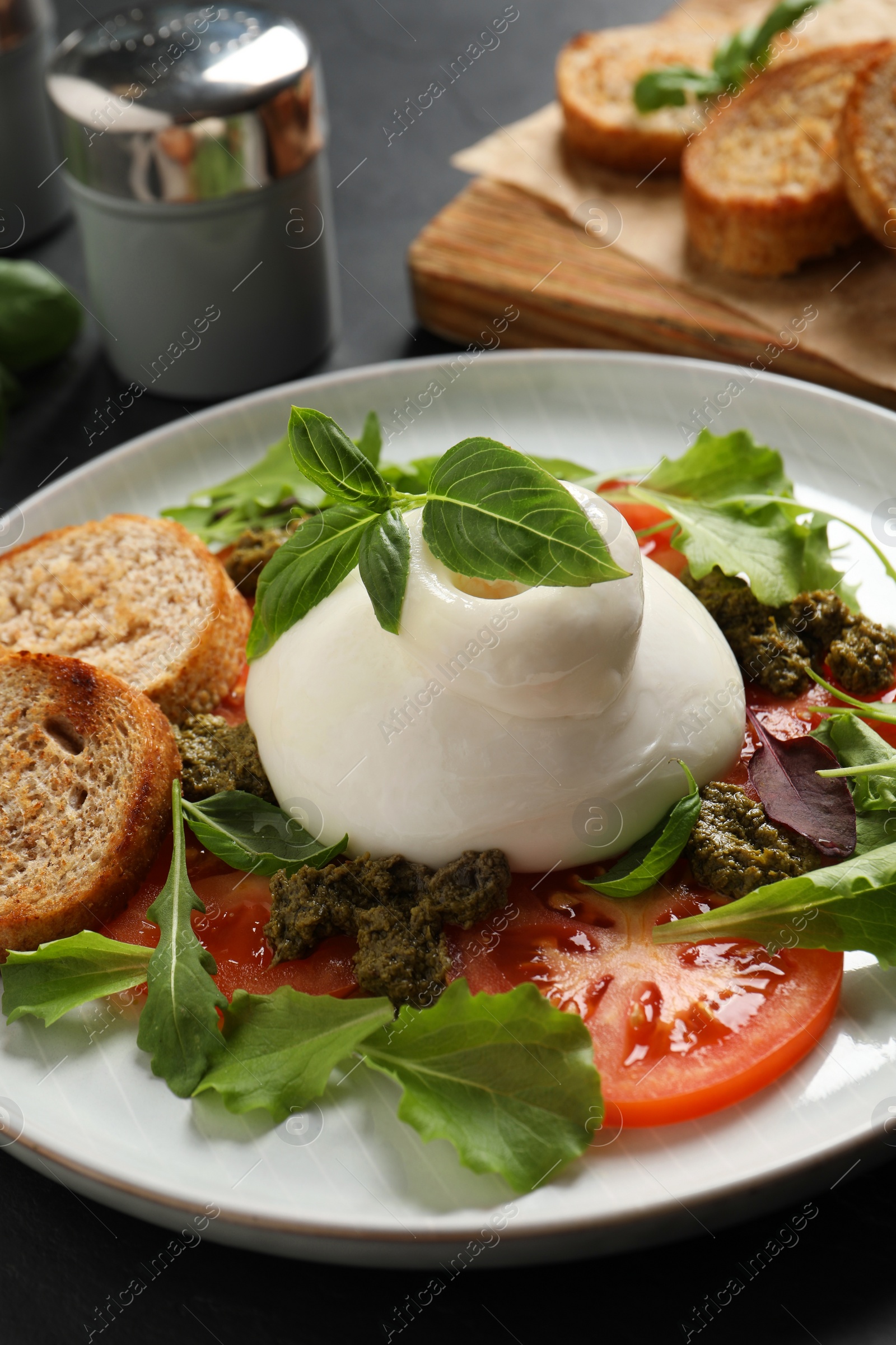 Photo of Delicious burrata salad with tomatoes, arugula and pesto sauce on dark grey table, closeup