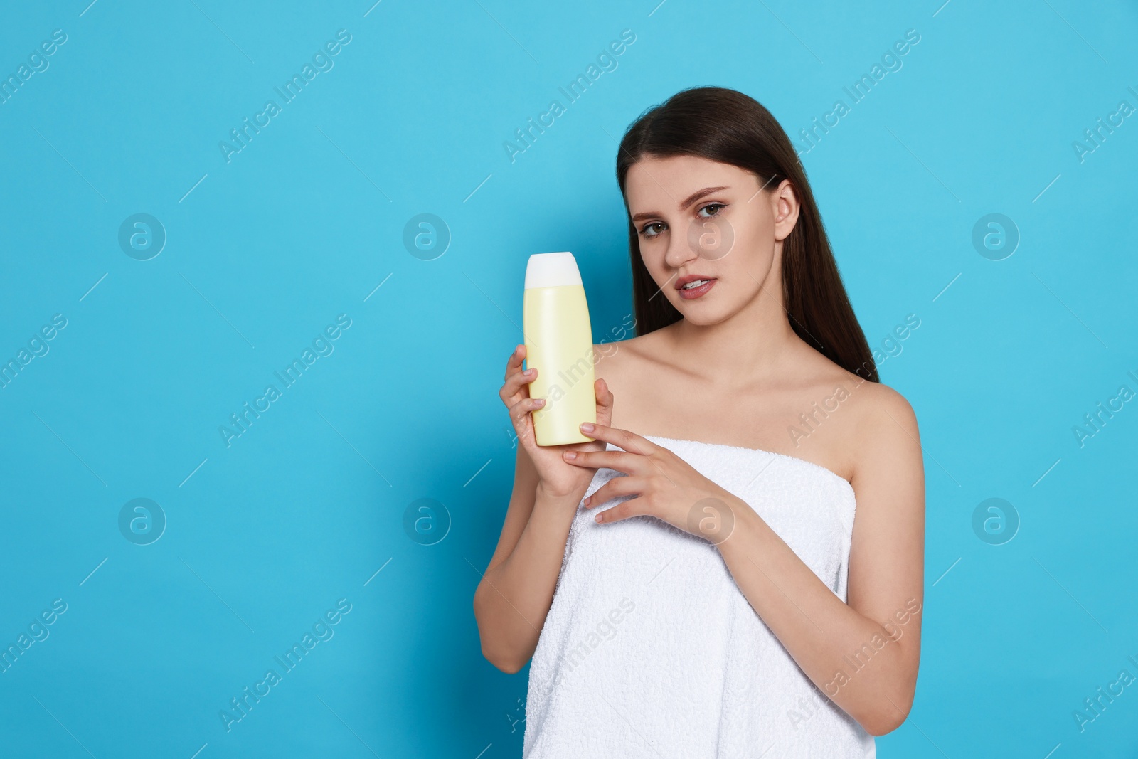 Photo of Beautiful young woman wrapped in towel holding bottle of shampoo on light blue background. Space for text