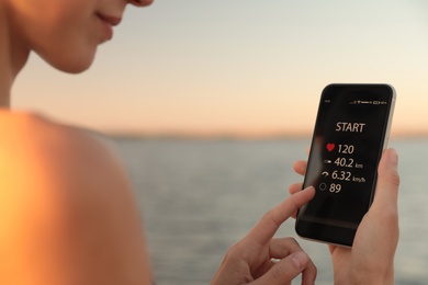 Photo of Young woman using fitness app on smartphone near river at sunset, closeup