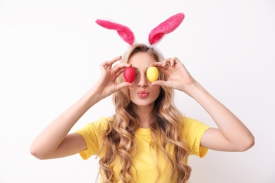 Photo of Beautiful young woman in bunny ears headband holding Easter eggs near eyes on light background