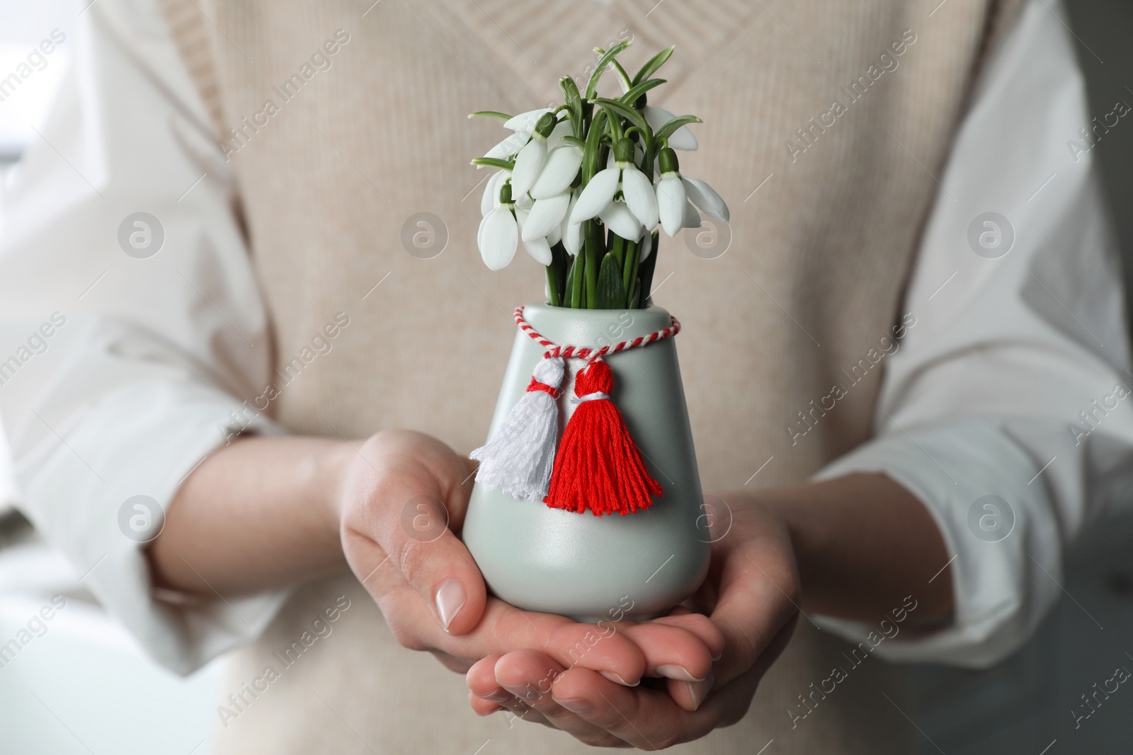 Photo of Woman holding vase with beautiful snowdrops and traditional martisor indoors, closeup. Symbol of first spring day