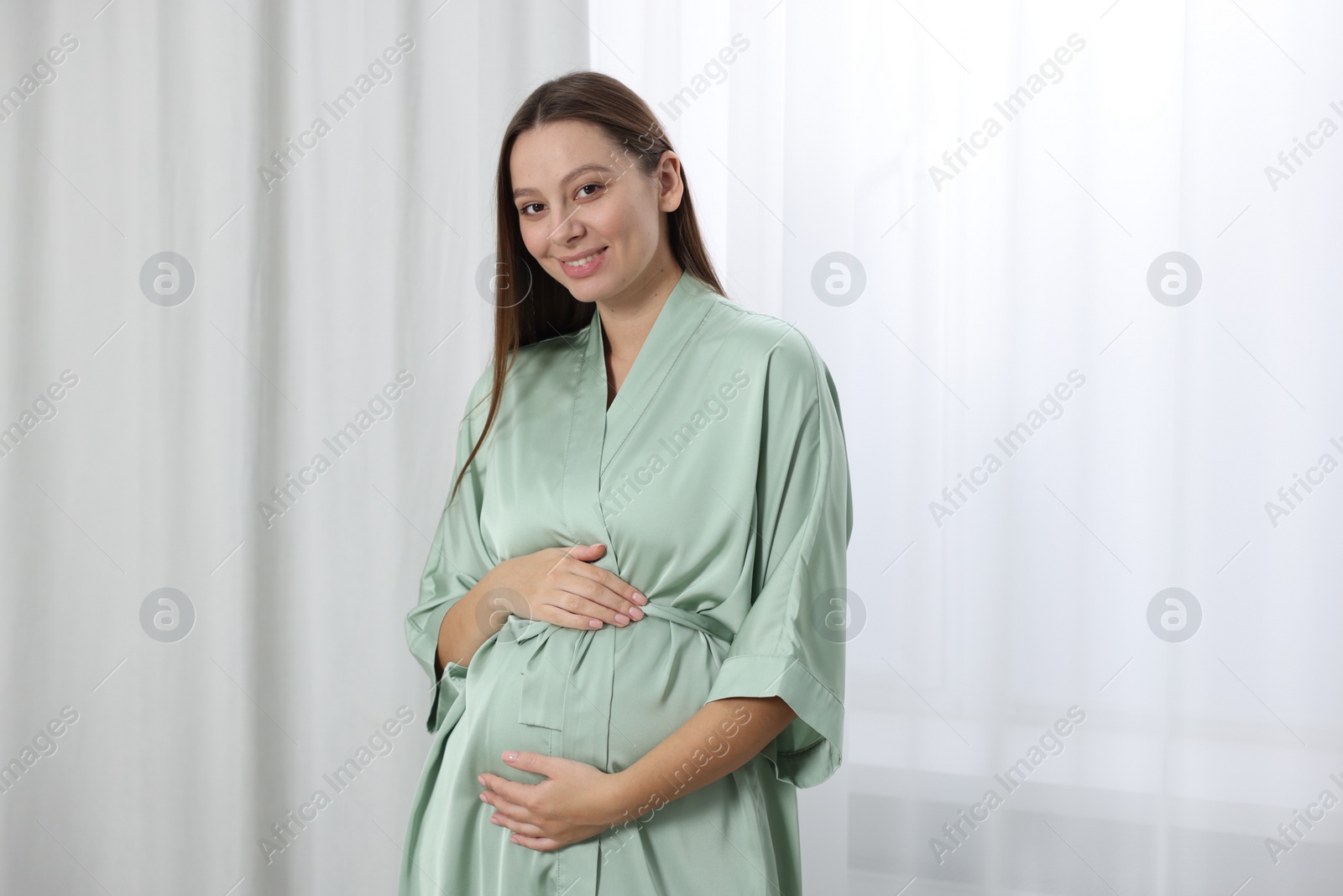 Photo of Beautiful pregnant woman in green dressing gown near window indoors