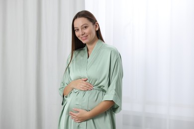 Photo of Beautiful pregnant woman in green dressing gown near window indoors