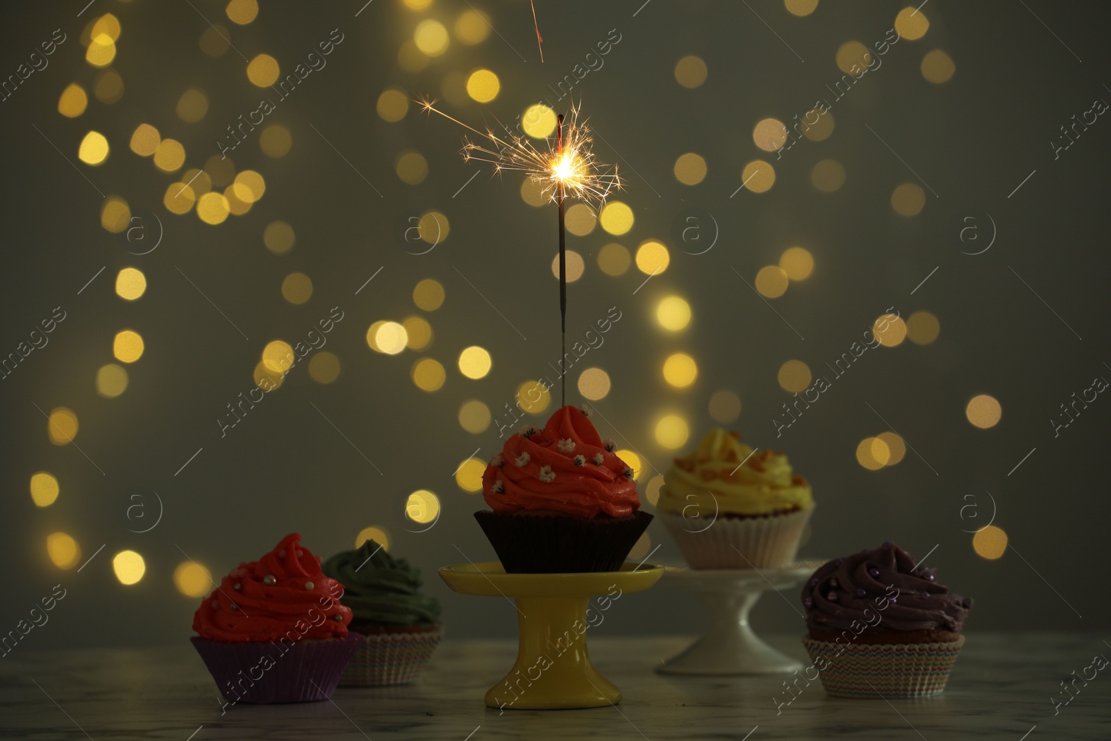 Photo of Different colorful cupcakes and one with sparkler on table against blurred lights