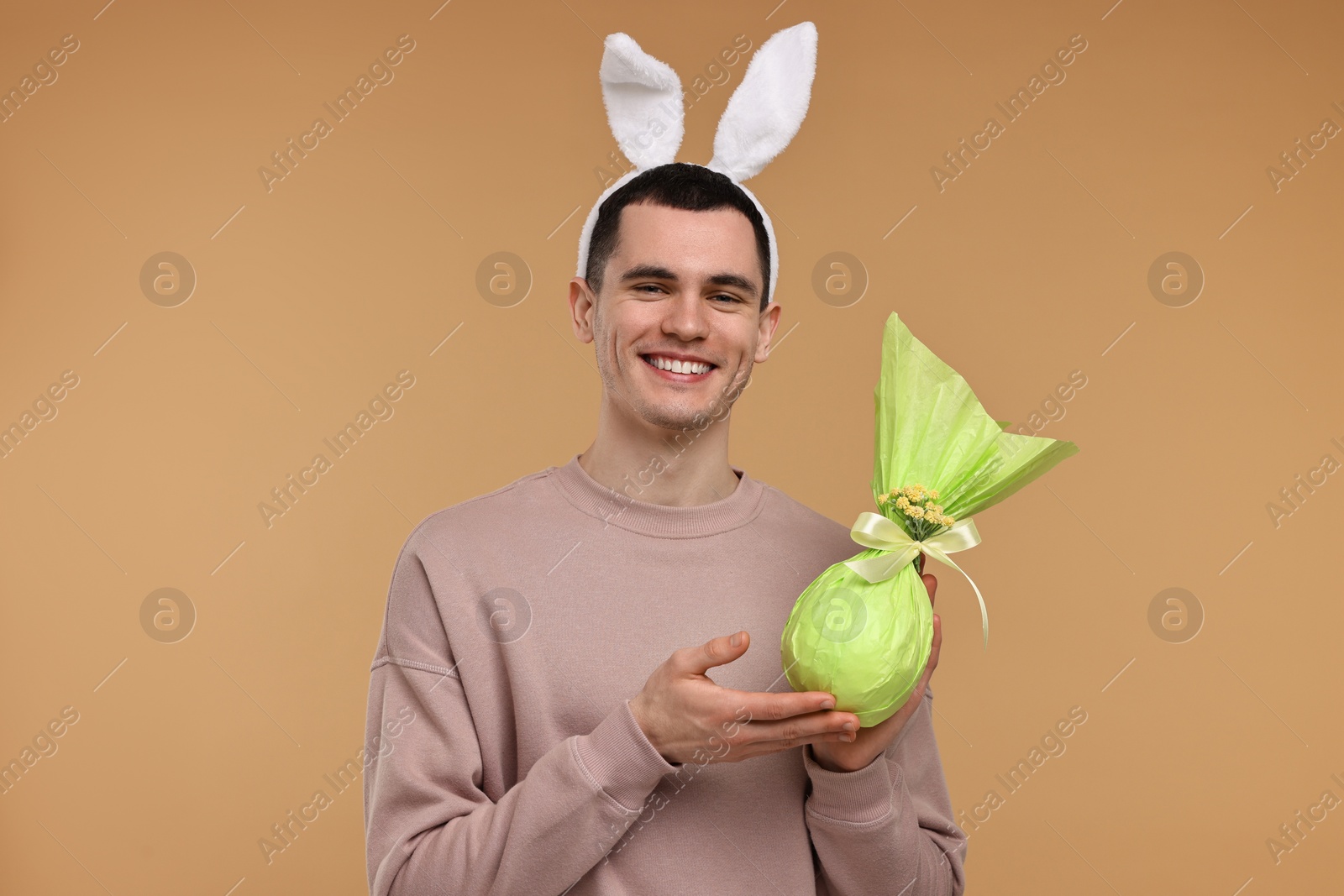 Photo of Easter celebration. Handsome young man with bunny ears holding wrapped gift on beige background