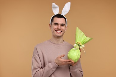 Photo of Easter celebration. Handsome young man with bunny ears holding wrapped gift on beige background