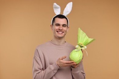 Easter celebration. Handsome young man with bunny ears holding wrapped gift on beige background