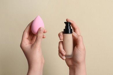 Photo of Woman holding bottle with skin foundation and makeup sponge on beige background, closeup