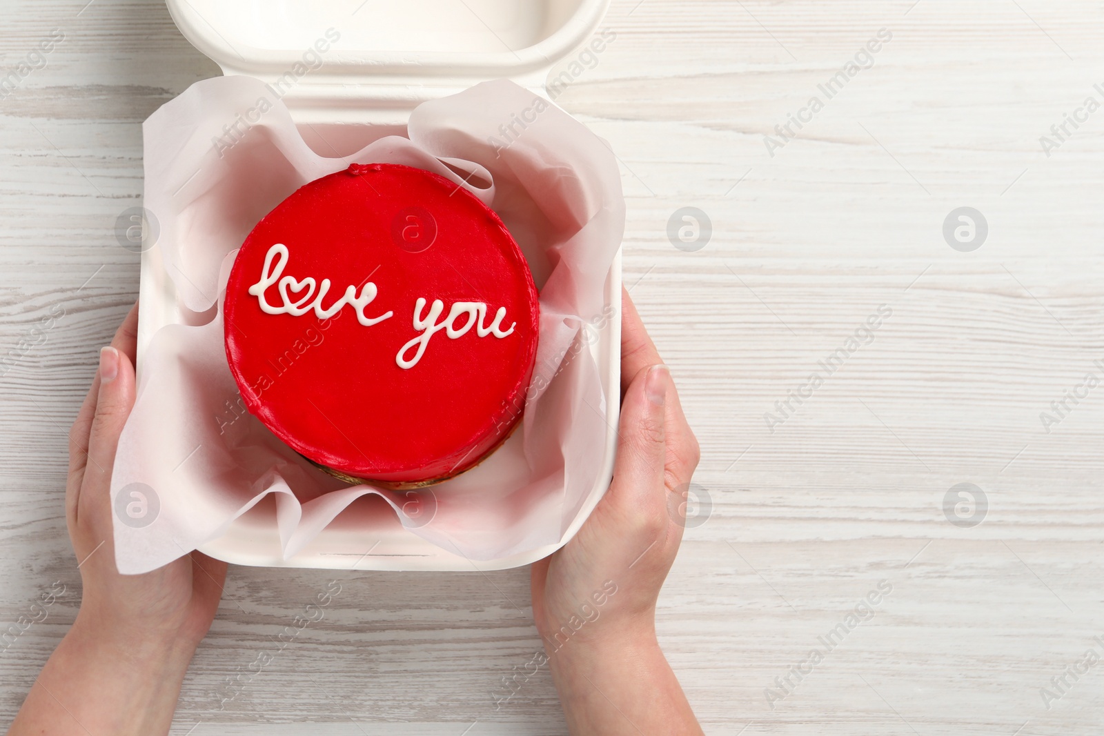 Photo of Woman holding takeaway box with bento cake at white wooden table, top view. St. Valentine's day surprise