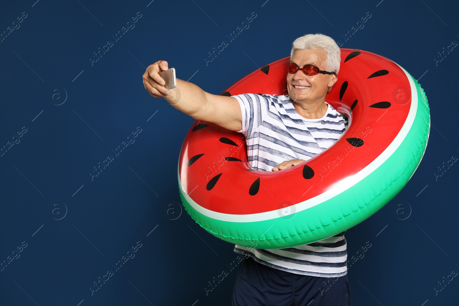 Photo of Funny mature man with bright inflatable ring taking selfie on dark blue background, space for text