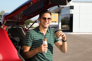 Young man with hot dog drinking coffee near car at gas station