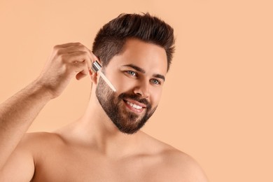 Handsome man applying cosmetic serum onto face on beige background, space for text