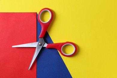 Photo of Red scissors and color sheets of paper on yellow background, top view. Space for text