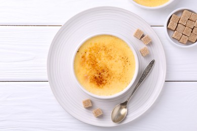 Delicious creme brulee in bowl, sugar cubes and spoon on white wooden table, top view