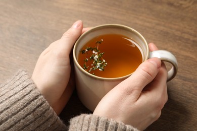 Photo of Woman holding cup of tasty herbal tea with thyme at wooden table, closeup