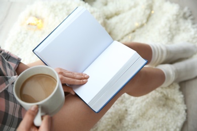 Photo of Young woman with cup of coffee reading book at home, above view