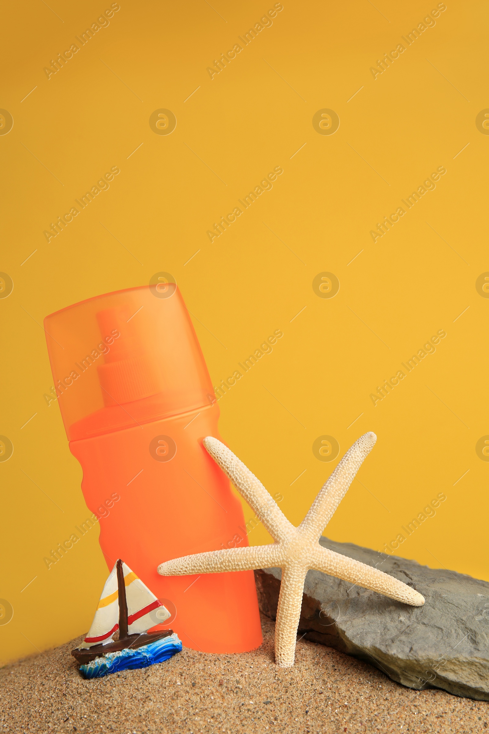 Photo of Sand with bottle of sunscreen, starfish, stone and ship figure against orange background. Sun protection