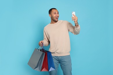 Happy African American man with shopping bags and smartphone on light blue background