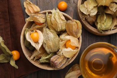 Ripe physalis fruits with calyxes in bowls and cup of tea on wooden table, flat lay