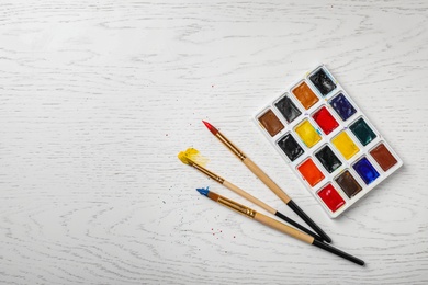 Plastic palette with colorful paints and brushes on wooden background, top view