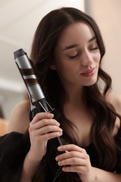 Beautiful woman using curling hair iron indoors