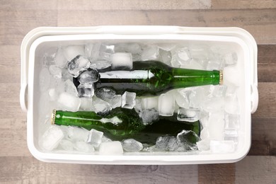 Plastic cool box with ice cubes and beer on wooden floor, top view