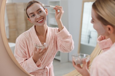 Photo of Woman applying face mask near mirror in bathroom. Spa treatments