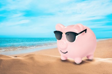 Vacation savings. Piggy bank with sunglasses on sandy beach near sea. Space for text