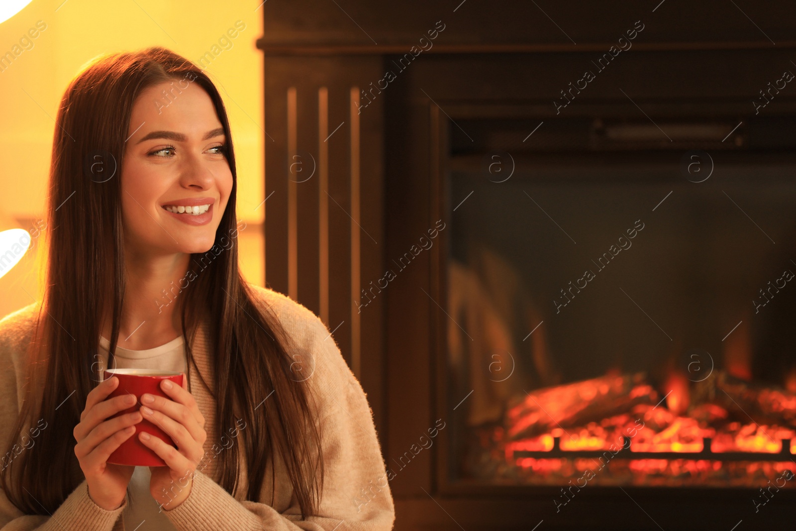 Photo of Young woman with cup of hot drink near fireplace indoors. Cozy atmosphere