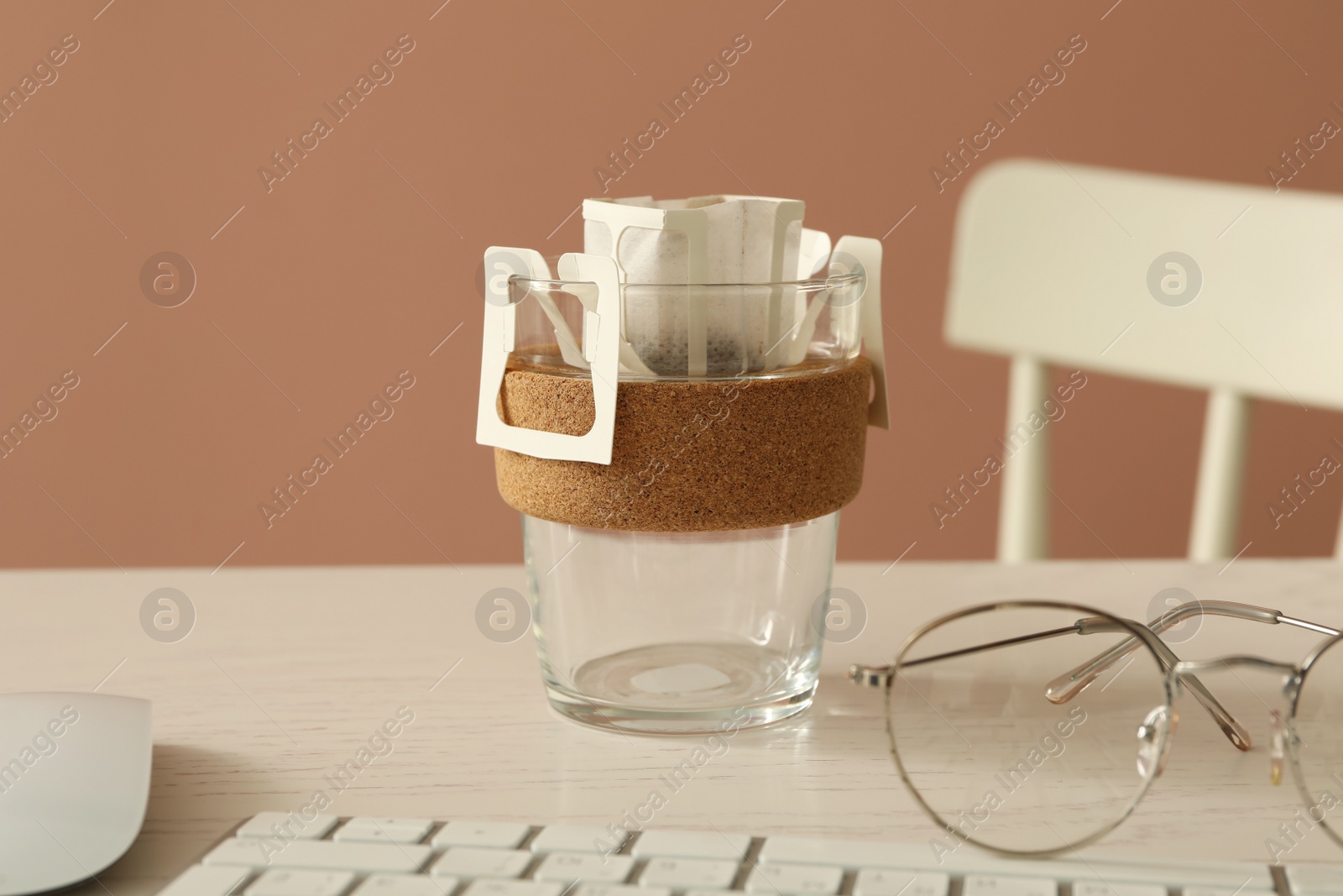 Photo of Glass cup with drip coffee bag, keyboard and glasses on white table, closeup