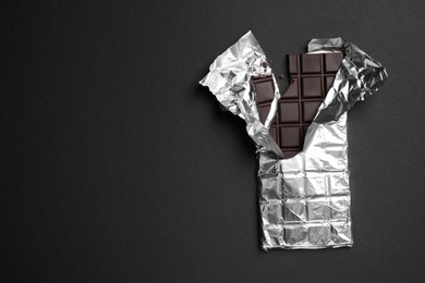 Photo of Broken dark chocolate bar wrapped in foil on black background, top view. Space for text