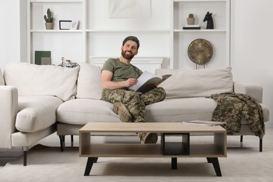 Photo of Happy soldier reading magazine on sofa in living room. Military service