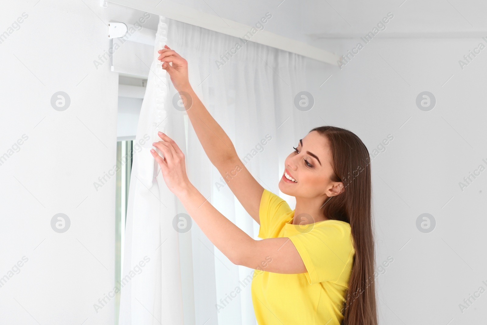 Photo of Young pretty woman hanging window curtain in room