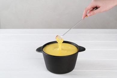 Woman dipping piece of bread into fondue pot with tasty melted cheese at white wooden table, closeup