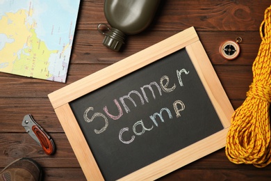 Chalkboard with text SUMMER CAMP and camping equipment on wooden background, flat lay