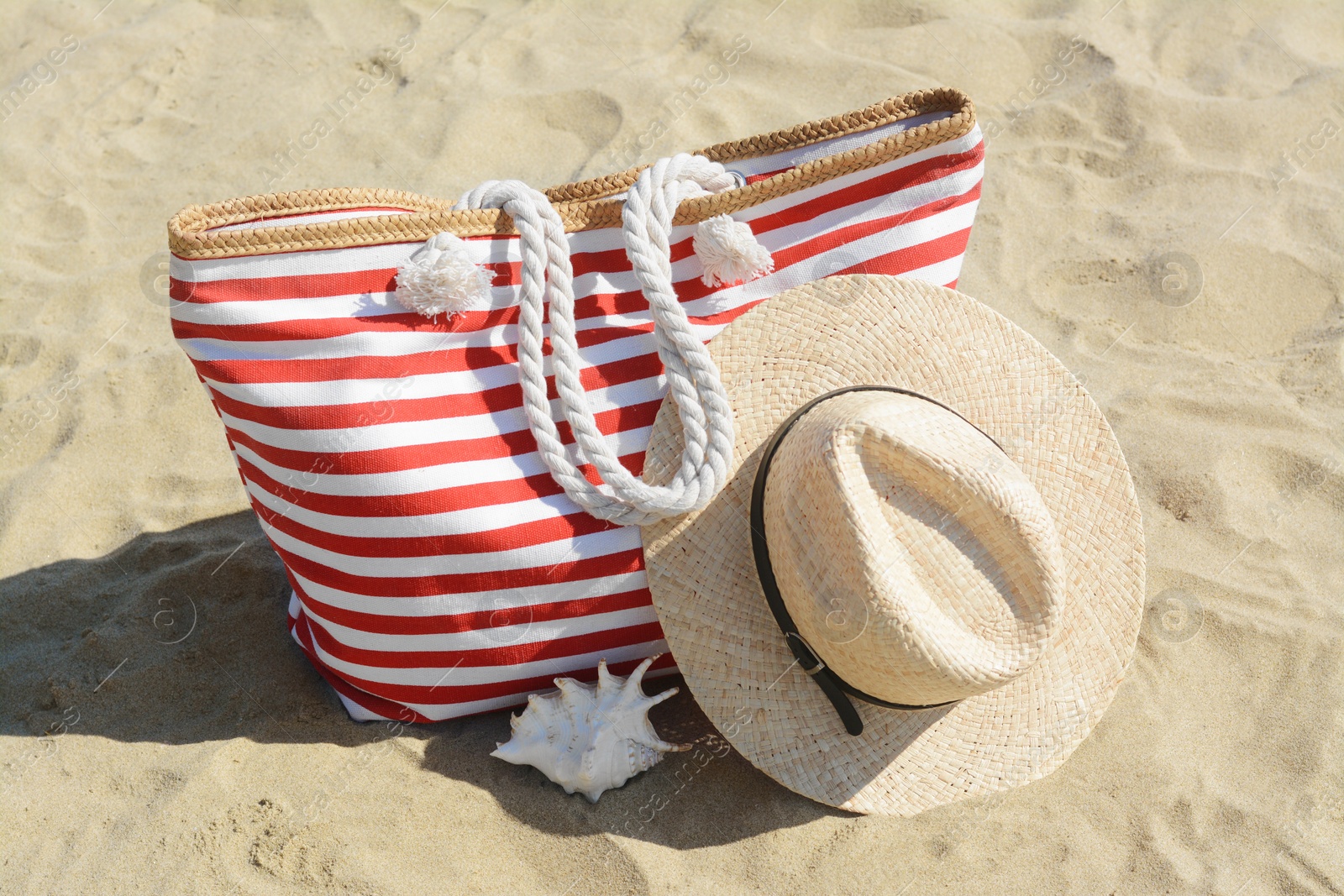 Photo of Stylish striped bag with straw hat and seashell on sandy beach