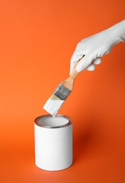 Person dipping brush into can of white paint on orange background