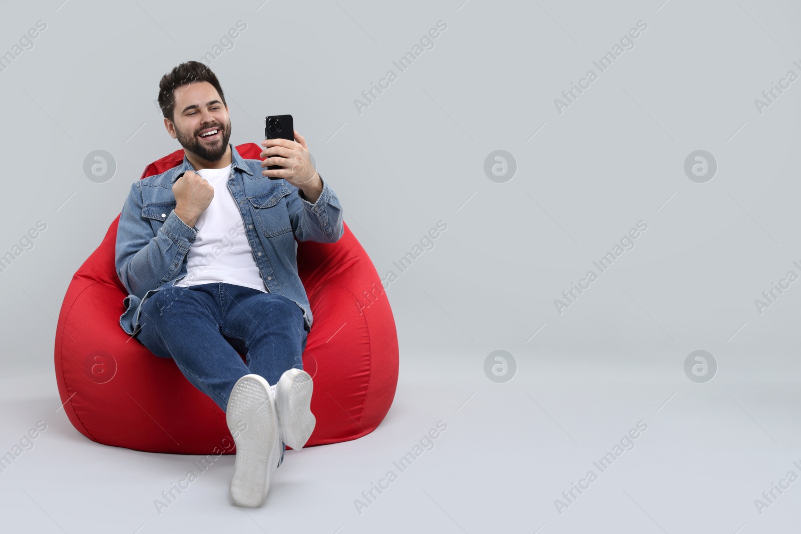 Photo of Happy young man using smartphone on bean bag chair against grey background, space for text