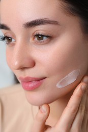 Photo of Young woman with dry skin applying cream onto her face on blurred background, closeup