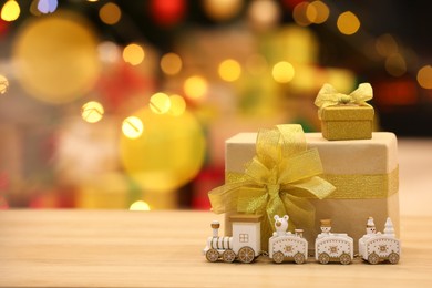 Photo of Beautiful gift boxes with toy train on wooden table against blurred festive lights, space for tex. Christmas celebration