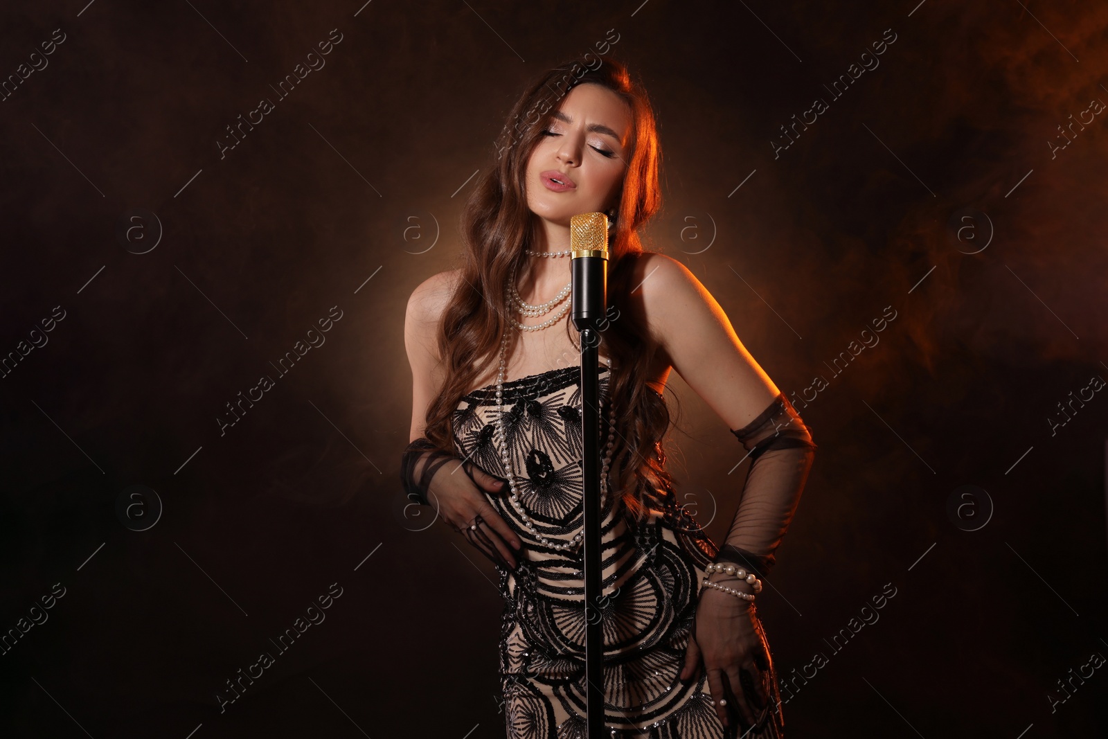 Photo of Beautiful young woman in stylish dress with microphone singing on dark background with smoke