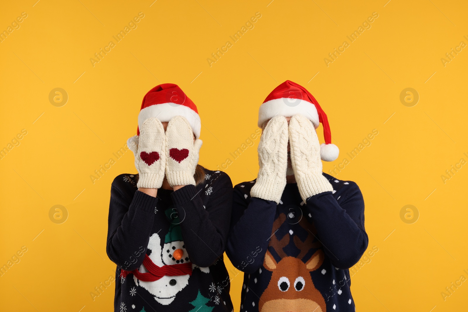 Photo of Couple in Christmas sweaters and Santa hats covering faces with hands in knitted mittens on orange background