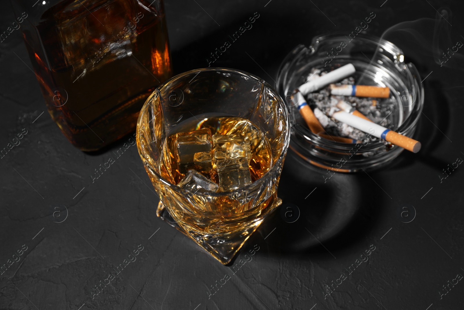 Photo of Alcohol addiction. Whiskey with ice cubes, smoldering cigarettes and ashtray on dark textured table, above view